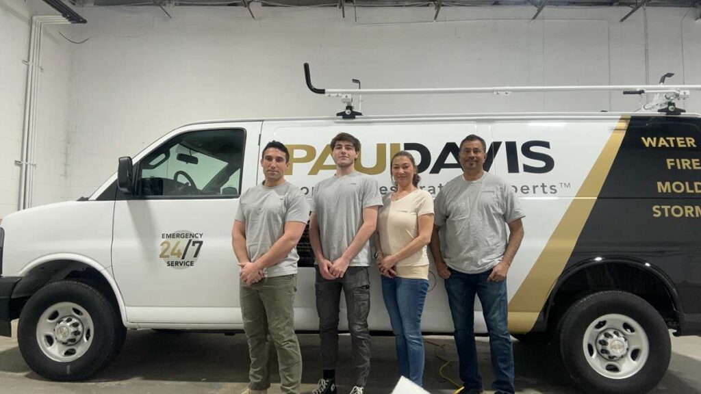 Team of four Paul Davis Restoration professionals standing in front of a branded service van inside a garage, representing Paul Davis Restoration of Middlesex County, NJ, with visible “24/7 Emergency Service” and services for water, fire, mold, and storm damage.